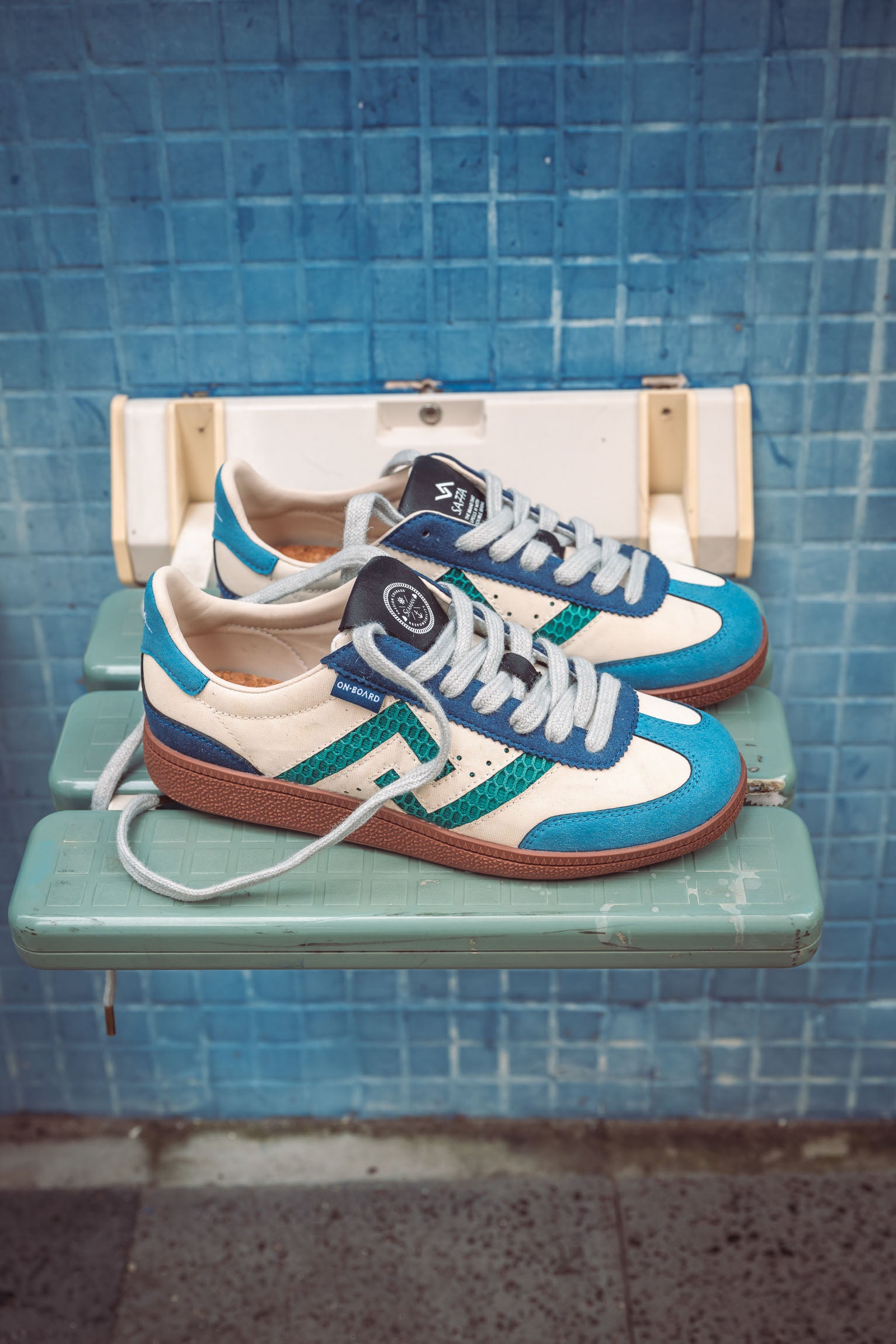 SA77A颯合_STEPS_YANG. The brand that spells H with double seven. Top ViewPair of blue and white sneakers with brown soles on a green shelf against a blue tiled wall.