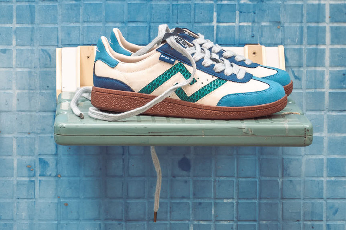 SA77A颯合_STEPS_YANG. The brand that spells H with double seven. Side ViewPair of blue and white sneakers with brown soles on a green shelf against a blue tiled wall.