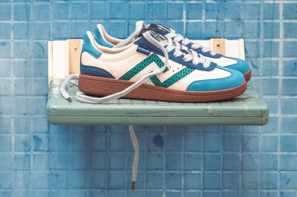 SA77A颯合_STEPS_YANG. The brand that spells H with double seven. Side ViewPair of blue and white sneakers with brown soles on a green shelf against a blue tiled wall.